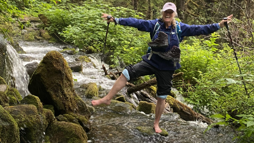 Toni barefoot with ski poles fording the stream, with a big smile on her face