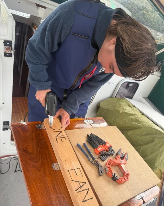 Crew member using a soldering iron to etch the name of s/v One Ocean on a board